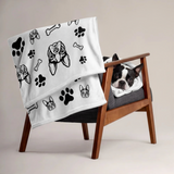 Dog lying on a wooden chair with a white blanket featuring black paw prints and Boston Terrier dog illustrations.