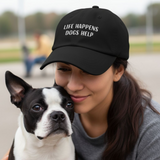 Woman wearing a black cap with 'Life Happens Dogs Help' text, holding a small black and white dog.